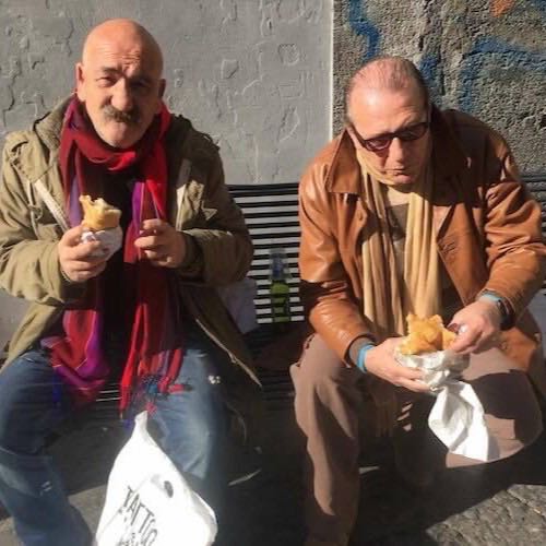 Two men on a bench in Naples eating pizza folded portafoglio style, in one hand